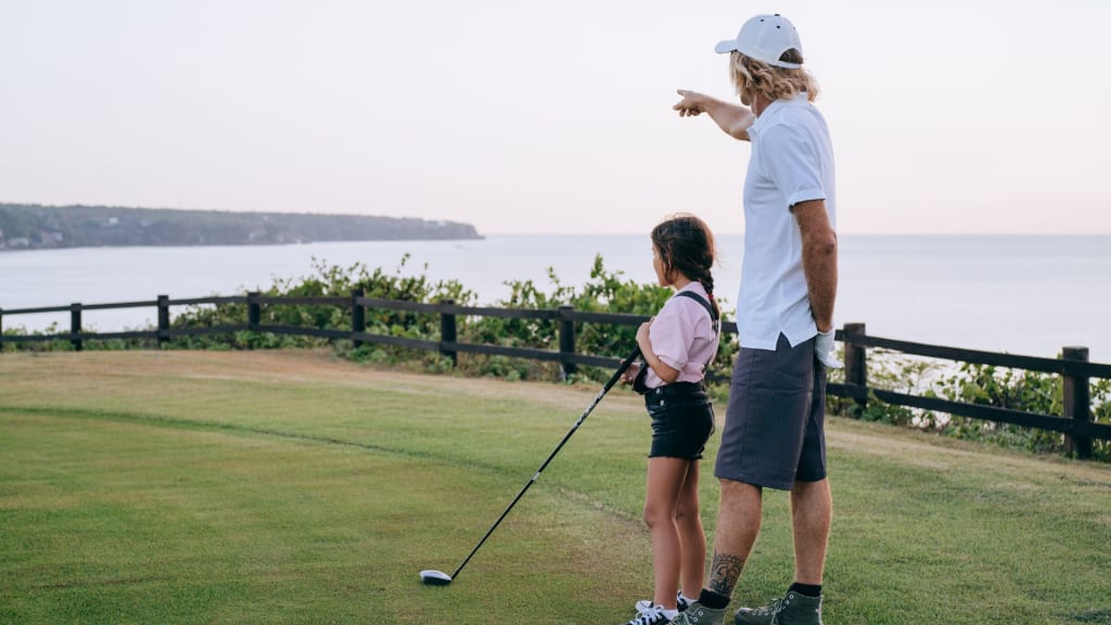 An adult and child stand on a grassy area near the ocean. The adult is pointing towards the horizon while the child holds a golf club.